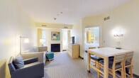 This thoughtfully designed hotel suite offers a comfortable and functional layout across a single floor, making it well-suited for extended stays or group travel. The living area serves as the heart of the suite, featuring a plush sectional sofa, a teal ottoman, a wall-mounted TV, and a cozy fireplace framed by colorful modern artwork. Natural light filters in through curtained windows, contributing to the suite's warm, inviting ambiance with soft yellow and cream tones throughout.

Adjacent to the living area is a dining space with a white-topped table and seating for four, flowing seamlessly into a fully equipped kitchen. The kitchen is outfitted with stainless steel appliances including a full-size refrigerator, oven, microwave, and dishwasher, along with ample cabinetry — ideal for preparing meals in-suite.

The suite includes two bedrooms, each furnished with a king-size bed, light wood headboard, coordinating nightstands, and soft blue accent walls that lend a calm, restful atmosphere. One bedroom features a wall-mounted TV and mirrored closet doors, while both offer natural light from exterior-facing windows.

Two bathrooms serve the suite: one featuring a clean vanity with a wide mirror and warm lighting, and the other with a full tub-shower combination with tile flooring — both finished in crisp white for a clean, polished look.