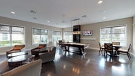 This single-floor amenity space is designed as a contemporary clubhouse lounge, offering a well-appointed environment for recreation and socializing. Rich, dark hardwood flooring runs throughout, providing a polished contrast to the warm neutral-toned walls and bright white trim. Recessed lighting and large roller-shade windows flood the interior with natural light while framing views of the outdoor pool and surrounding grounds.

The main lounge area features a generous arrangement of plush swivel chairs and sofas grouped around mosaic-style accent tables, complemented by a wall-mounted flat-screen TV set atop a textured stone media console. Colorful modern artwork adorns the walls, adding visual interest to the space.

At the center of the room, a full-size billiards table commands attention beneath a sleek industrial-style pendant light fixture, making it a natural focal point for entertaining. A wall-mounted television above the billiards area adds to the entertainment appeal.

A dedicated dining nook is situated in a windowed corner, bathed in natural light from expansive corner windows that offer direct views of the pool area. A round table with upholstered chairs provides an intimate setting for casual gatherings. Glass-panel doors lead to an adjacent room, suggesting additional amenity or office space, further enhancing the versatility of this well-designed communal facility.