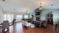 Located in O'Fallon, Illinois, this single-story clubhouse at Fairfield Place offers a welcoming and well-appointed gathering space for residents. The expansive main lounge is the heart of the clubhouse, anchored by a dramatic floor-to-ceiling stacked stone fireplace with a wood mantel and wall-mounted television above. Vaulted ceilings rise over the seating area, lending an open, airy feel, while rich hardwood flooring runs throughout the entire space. Stylish orb pendant chandeliers hang overhead, adding warmth and character to the room. Comfortable leather sofas and upholstered chairs are arranged to encourage conversation and relaxation. A cozy nook near large windows fitted with decorative valances and blinds provides views of the surrounding neighborhood and features high-top bistro seating—ideal for casual gatherings. The full kitchen is equipped with oak cabinetry, granite countertops, a mosaic tile backsplash, and black appliances including a range, microwave, and refrigerator. A water cooler station and a pass-through to the main lounge add practical convenience. The overall ambiance blends rustic charm with comfortable, communal living, making this clubhouse a functional and attractive amenity for the Fairfield Place community.