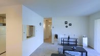 This single-floor space is designed with a clean, minimalist aesthetic, characterized by bright white walls and neutral-toned carpeting that create a light and airy atmosphere throughout. The main living area is open and uncluttered, offering a comfortable sense of space. A low-profile media console serves as a functional focal point along one wall, accompanied by decorative framed artwork that adds subtle visual interest to the otherwise understated décor.

A wall-mounted mirror with a louvered frame enhances the sense of openness, reflecting light and adding depth to the room. Small decorative accents, including single-stem flowers in slender vases, contribute to the tidy, simple styling of the space.

A central hallway connects the living area to the rest of the unit, with doorways visible in the distance suggesting access to additional rooms. The corridor is well-lit, maintaining the bright and neutral character found throughout the space. The overall ambiance is calm and functional, with a straightforward layout that prioritizes ease of movement and practicality. The consistent use of light colors across walls, flooring, and furnishings contributes to a cohesive and welcoming interior environment.