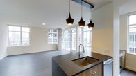 This single-floor space showcases a contemporary open-concept design that integrates the kitchen and living areas into one cohesive, light-filled environment. The kitchen centers around a substantial dark-topped island equipped with a deep undermount sink finished in a warm gold tone and a pull-down faucet, complemented by warm wood cabinetry below. Three matte black pendant lights hang above the island in varying sizes, adding a stylish focal point overhead. Recessed lighting supplements the natural light that pours in through the expansive floor-to-ceiling windows lining the perimeter, framing views of the surrounding urban streetscape and a private outdoor balcony area. The living space extends generously beyond the kitchen, highlighted by smooth, wide-plank gray flooring that runs throughout and anchors the neutral color palette of white walls and light ceilings. A structural concrete column near the living area adds an industrial design element that contrasts subtly with the otherwise refined interior finishes. The overall ambiance is clean, bright, and modern, well-suited for both everyday living and entertaining.