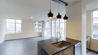 This single-floor space showcases a contemporary open-concept design that integrates the kitchen and living areas into one cohesive, light-filled environment. The kitchen centers around a substantial dark-topped island equipped with a deep undermount sink finished in a warm gold tone and a pull-down faucet, complemented by warm wood cabinetry below. Three matte black pendant lights hang above the island in varying sizes, adding a stylish focal point overhead. Recessed lighting supplements the natural light that pours in through the expansive floor-to-ceiling windows lining the perimeter, framing views of the surrounding urban streetscape and a private outdoor balcony area. The living space extends generously beyond the kitchen, highlighted by smooth, wide-plank gray flooring that runs throughout and anchors the neutral color palette of white walls and light ceilings. A structural concrete column near the living area adds an industrial design element that contrasts subtly with the otherwise refined interior finishes. The overall ambiance is clean, bright, and modern, well-suited for both everyday living and entertaining.