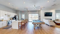 Located in Carolina Shores, NC, this single-story community clubhouse offers a well-appointed gathering space designed for both relaxation and activity. The open-concept main area features warm light hardwood flooring throughout, large ceiling fans, and a striking glass garage door that floods the space with natural light while providing views of the surrounding neighborhood. A shiplap fireplace anchors the comfortable lounge area, which is outfitted with plush seating and a wall-mounted TV, creating an inviting social hub. Adjacent to the lounge, a full-size pool table adds a recreational focal point to the entertaining space. The fully equipped kitchen is a standout feature, boasting white shaker cabinetry, quartz countertops, a subway tile backsplash, stainless steel appliances, and a large island with bar seating — ideal for hosting community events. A separate dining area accommodates multiple round tables and chairs, bathed in natural light from several windows with vaulted ceilings adding to the airy feel. The dedicated fitness center is well-equipped with strength-training machines, cardio equipment including treadmills, resistance stations, and a wall-mounted TV, all set within a bright, ventilated room. Soft gray walls and consistent hardwood-style flooring unify the entire space with a clean, coastal aesthetic.