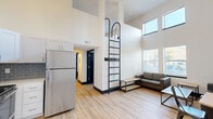 Located at 221 E State St in West Lafayette, Indiana, this 2-bedroom, 2-bathroom unit delivers a striking architectural design that maximizes vertical space. The open-concept living and kitchen area is anchored by soaring double-height ceilings, with tall windows flooding the space with natural light and offering views of the surrounding neighborhood. A sleek black metal loft ladder rises dramatically from the living area, serving as both a functional and visual centerpiece.

The kitchen is outfitted with a full suite of stainless steel appliances — including a refrigerator, range, microwave, and dishwasher — complemented by white shaker cabinetry, gray subway tile backsplash, and light hardwood-style flooring that extends throughout the unit. The living area provides ample room for seating and a dining setup beneath the expansive ceiling volume.

Both bedrooms are compact yet functional, each featuring hardwood-style floors and built-in or standard furniture to optimize the available space. The hallway connecting the rooms includes additional cabinetry for storage. Two full bathrooms serve the unit, providing practical convenience for multiple occupants. A dedicated closet adds further storage capacity. The clean, modern finishes and efficient layout make this unit a well-designed space in the heart of West Lafayette.
