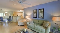 This single-floor residence offers a warm and inviting atmosphere with a thoughtfully designed open-concept layout. The living area serves as the heart of the space, featuring light hardwood-style flooring, a ceiling fan overhead, and a soothing color palette highlighted by a striking periwinkle blue accent wall. Framed palm tree artwork adds a coastal, tropical touch that carries throughout the room's décor. The living room is comfortably appointed with a sage green sofa and coordinating patterned armchair, centered around a glass-topped coffee table that enhances the sense of openness and light. A warm table lamp provides ambient lighting, complementing the natural light that fills the space. Flowing seamlessly from the living area is a functional kitchen, visible through an open pass-through bar counter. The kitchen features light wood cabinetry, under-cabinet lighting, and clean white appliances, creating a bright and efficient cooking space. The bar-height counter serves as a natural dividing element between the living and kitchen areas, ideal for casual dining or entertaining. Overall, the space presents a relaxed, coastal-inspired ambiance with a clean, well-maintained interior that is both functional and aesthetically cohesive.