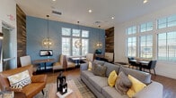 Located at 7210 Reading Rd in Rosenberg, Texas, this single-floor community amenity space offers a well-appointed gathering area designed for both relaxation and social interaction. The open layout is anchored by a comfortable lounge zone furnished with a plush gray sofa, accent pillows in yellow and gray, and a cognac leather armchair, all arranged atop a textured area rug over warm hardwood-style flooring.

A series of built-in dining booths line the perimeter, each paired with a wall-mounted flat-screen TV and illuminated by striking globe pendant lights in a brushed gold finish. Large windows with white plantation shutters flood the space with natural light and offer views of the surrounding outdoor greenery. The soaring ceiling height enhances the open, airy feel of the room.

One wall features a dramatic reclaimed wood accent panel that adds warmth and texture, serving as a focal point throughout the space. Adjacent to the main lounge is a sleek, fully equipped kitchenette outfitted with stainless steel appliances—including a refrigerator, wall oven, and undermount sink—set beneath white countertops against a matching wood-panel backdrop. The overall design balances contemporary aesthetics with inviting comfort, making this a functional and visually compelling communal space.