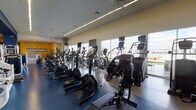 Located at 503 George Bush Dr W in College Station, Texas, this fitness center is a comprehensive, single-floor facility designed to support a wide range of workout needs. The space is organized into distinct zones that flow seamlessly into one another, creating an open and energizing environment throughout.

The cardio area is a standout feature, lined with rows of treadmills, ellipticals, stair climbers, and stationary bikes from leading equipment brands. Floor-to-ceiling windows along the perimeter flood the space with natural light and offer expansive outdoor views, enhancing the workout experience. Ceiling fans contribute to a comfortable atmosphere in the cardio zones.

The strength training section provides a thorough selection of selectorized machines, free weights, cable systems, and functional training equipment, all arranged to allow for efficient use of the floor space. Mirrors along the walls support proper form and add depth to the room.

A dedicated open-floor studio area accommodates group fitness activities and functional training, outfitted with specialty equipment and colorful, motivational wall graphics. The entrance corridor features vibrant artwork, a water station, and a front desk, establishing a welcoming arrival experience. The color palette throughout — incorporating bold yellows, blues, and greens — contributes to the facility's dynamic and modern character.