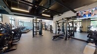 Located at 174 N Addison Ave in Elmhurst, Illinois, this single-floor fitness center is designed to support a comprehensive workout experience across clearly defined training zones. The space features an industrial-inspired aesthetic, with exposed black structural beams and ductwork contrasting against white walls and durable rubber flooring throughout.

The strength training area is generously proportioned, housing multiple cable machine systems, weight benches, and a row of free-weight and functional training equipment. Full-length mirrors line one wall, enhancing the sense of space and allowing users to monitor their form. Linear pendant lighting suspended from the exposed ceiling framework provides consistent, focused illumination across this zone.

A dedicated cardio section includes a row of treadmills positioned facing large floor-to-ceiling windows, allowing natural light to fill the room and offering views of the surrounding streetscape. A ceiling fan overhead contributes to ventilation and comfort during use.

A separate area accommodates stationary bikes and cycling equipment, also benefiting from tall windows and direct access to the outdoors. Near the entrance, Matrix-brand selectorized strength machines are positioned alongside a decorative wall mural, creating a welcoming transition into the facility. The overall ambiance is modern, functional, and well-maintained.