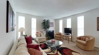 This single-floor space centers around a well-appointed living room that demonstrates a practical and welcoming design. The room benefits from an abundance of natural light, courtesy of multiple tall, narrow windows fitted with white blinds positioned along the walls, creating an airy and open atmosphere throughout.

The living room features neutral-toned walls and a textured ceiling that complement the light carpeted flooring. A fireplace serves as a subtle focal point along one wall, adding a cozy architectural element to the space. The room is generously proportioned, accommodating a large sectional sofa dressed with red and cream accent pillows, a glass-top coffee table with ornate metalwork legs, and additional seating in the form of cushioned armchairs.

A bold patterned area rug in shades of red, brown, black, and cream anchors the seating arrangement and adds visual warmth to the neutral palette. A tall indoor tree plant introduces a natural element, enhancing the room's inviting character. Entertainment is served by a flat-screen television positioned on a wicker-style media stand in the corner. Overall, the space presents a comfortable, casually elegant living environment with thoughtful attention to both function and style.