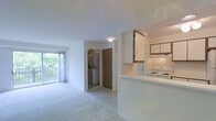 This single-floor unit features a thoughtfully arranged layout that maximizes functionality within a well-proportioned footprint. The open living area is finished with neutral wall-to-wall carpeting, creating a warm and inviting atmosphere, while tile flooring defines the entryway and transitional spaces. Natural light flows into the living space through a sliding glass door that leads to a private balcony, offering outdoor access and views of surrounding greenery.

The kitchen is positioned adjacent to the living area and features a pass-through counter that connects the two spaces, promoting an open feel. White cabinetry with wood-accented upper cabinets provides ample storage, and white appliances including a range are included. A dedicated utility area accommodates in-unit washer and dryer connections, adding notable convenience.

A wood-toned closet door is visible from the main living space, providing generous storage. The entry foyer, framed by a white front door, offers a clean transition into the unit. The overall design presents a clean, neutral palette throughout, making the space move-in ready and easy to personalize.