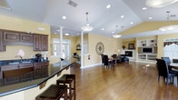 Claysville Landing offers a thoughtfully designed single-story community space that combines functionality with a warm, inviting atmosphere. The open-concept layout allows for a seamless flow throughout the interior, anchored by rich hardwood flooring that runs the length of the space.

The vaulted ceiling adds a sense of height and openness, complemented by a combination of recessed lighting and elegant pendant fixtures that provide ample illumination throughout. Decorative white columns help subtly define distinct areas without interrupting the open feel of the room.

A dark-granite countertop bar area with rich wood cabinetry is situated toward the front, offering a practical and polished focal point. Large glass-paned French doors allow generous natural light to fill the interior while providing views of the outdoors. A dedicated workspace or business center area is visible near the entrance, adding convenience for residents.

Toward the rear, a dining area with dark wood furniture sits adjacent to a cozy lounge space featuring a fireplace with a classic white surround, ideal for relaxed gatherings. Warm yellow walls throughout contribute to the cheerful, welcoming ambiance of the entire space.