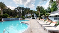 This outdoor pool area offers a well-appointed resort-style retreat designed for relaxation and leisure. The centerpiece is a generously sized freeform swimming pool with crystal-clear blue water, framed by a decorative tiled accent wall that adds a distinctive architectural touch. The pool's curved design creates an inviting, organic shape that enhances the overall aesthetic of the space.

Surrounding the pool is a broad, paver-lined sun deck that provides ample space for lounging. A row of cushioned chaise lounge chairs lines one side of the deck, offering comfortable seating for sunbathing or unwinding poolside. Additional seating and shaded areas with umbrellas are available for guests who prefer to relax out of direct sunlight.

The space is beautifully landscaped with tall, swaying palm trees, manicured shrubs, and lush greenery that create a tropical ambiance. The mature trees provide a natural backdrop that enhances privacy and adds to the serene atmosphere. Residential buildings are visible beyond the treeline, situating the pool area as a central amenity within the community.

Open to the sky, the pool area benefits from abundant natural light throughout the day, making it an ideal outdoor gathering and recreation space for residents and guests alike.