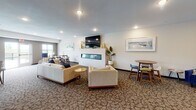 Located at 540 Tyler Rd S in Red Wing, Minnesota, this single-floor community common space is thoughtfully designed to encourage social interaction and comfortable everyday living. The layout flows seamlessly across distinct functional zones, all unified by patterned carpeting, recessed lighting, and a warm, contemporary aesthetic.

The fully equipped communal kitchen serves as a central hub, featuring rich dark cabinetry, granite countertops, stainless steel appliances including a refrigerator, range, microwave, and dishwasher, and a generous island with bar-height seating. Pendant lighting above the island adds a refined touch, while light wood-look tile flooring grounds the space practically and stylishly.

Adjacent to the kitchen, a coffee station with a single-serve brewer and dark wood credenza provides a convenient spot for daily refreshment. Decorative gold ginkgo leaf wall art adds an artistic accent to this transitional area.

The lounge area is expansive and inviting, anchored by a sleek linear fireplace framed in white tile with a wall-mounted television above. Comfortable neutral sofas and accent chairs are arranged for both intimate conversation and open gathering. Multiple dining and café-style seating clusters are positioned near large windows, filling the space with natural light and offering views of the surrounding landscape. The overall ambiance is modern, relaxed, and well-suited for community life.