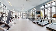 This modern fitness facility in Austin, TX offers a spacious, single-floor workout environment designed to support a wide range of exercise routines. The open-concept layout is bathed in natural light, courtesy of large, black-framed windows that line multiple walls and extend toward the elevated ceiling, creating an airy and energizing atmosphere. Ceiling fans provide additional airflow throughout the space, enhancing comfort during intense workouts.

The gym is comprehensively equipped to accommodate both cardio and strength training. The cardio zone features multiple treadmills, elliptical trainers, and a stair climber, each positioned to take advantage of the outdoor views. A wall-mounted flat-screen television is centrally placed for entertainment during workouts. The strength training area includes a full dumbbell rack with a wide range of weights, adjustable benches, a multi-functional cable machine with TRX suspension straps, a weight rack loaded with barbells, and a dedicated free weight bench press setup. Plyo boxes and additional functional training accessories round out the equipment selection.

The flooring throughout is durable rubber-textured material, well-suited for high-impact activity. Full-length mirrors along one wall amplify the sense of space and allow users to monitor their form. The clean, neutral color palette of soft grays and whites contributes to a bright, professional ambiance throughout this well-appointed fitness space.