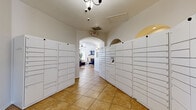 This single-floor community space is divided into two distinct functional areas that together create a welcoming and practical environment for residents. The package locker corridor serves as the first point of entry, lined on both sides with rows of white parcel lockers offering secure package management. The corridor is finished with warm terracotta-toned tile flooring and features an elegant arched passageway that leads into the main lounge area, adding a subtle architectural charm to the transition between spaces. Overhead, a chandelier and climate control vents contribute to a well-maintained atmosphere throughout.

The main lounge area is bright and thoughtfully designed, benefiting from multiple windows fitted with white blinds that allow natural light to filter in. A white fireplace serves as a central focal point, framed by a wall-mounted television and flanked by indoor plants that add warmth and life to the space. The seating arrangement includes a mix of navy blue accent chairs, tan leather armchairs, and a central coffee table set atop a richly patterned area rug. Framed landscape artwork and a wooden credenza further enhance the room's polished, contemporary aesthetic. A glass-paneled exterior door provides direct outdoor access, completing the functional and inviting character of this community gathering space.