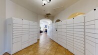 This single-floor community space is divided into two distinct functional areas that together create a welcoming and practical environment for residents. The package locker corridor serves as the first point of entry, lined on both sides with rows of white parcel lockers offering secure package management. The corridor is finished with warm terracotta-toned tile flooring and features an elegant arched passageway that leads into the main lounge area, adding a subtle architectural charm to the transition between spaces. Overhead, a chandelier and climate control vents contribute to a well-maintained atmosphere throughout.

The main lounge area is bright and thoughtfully designed, benefiting from multiple windows fitted with white blinds that allow natural light to filter in. A white fireplace serves as a central focal point, framed by a wall-mounted television and flanked by indoor plants that add warmth and life to the space. The seating arrangement includes a mix of navy blue accent chairs, tan leather armchairs, and a central coffee table set atop a richly patterned area rug. Framed landscape artwork and a wooden credenza further enhance the room's polished, contemporary aesthetic. A glass-paneled exterior door provides direct outdoor access, completing the functional and inviting character of this community gathering space.
