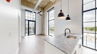 Located at 4715 West Irving Park Road in Chicago, IL, this industrial-chic loft blends raw architectural character with contemporary design. The open-concept living area commands attention with soaring ceilings, exposed concrete columns, and visible ductwork overhead — hallmarks of a thoughtfully converted industrial space. Towering steel-framed windows flood the interior with abundant natural light while offering views of the surrounding neighborhood, and a private balcony extends the living space outdoors.

The kitchen is appointed with striking veined granite countertops, white cabinetry, stainless steel appliances, and a matte black faucet. Bold pendant lighting in matte black adds a modern contrast to the bright, airy aesthetic. Light wood-tone flooring runs throughout, complementing the clean white walls and lending warmth to the industrial framework above.

Two hallways connect the living spaces efficiently, leading to two well-appointed bathrooms. Each bathroom features rich brown mosaic tile flooring with marble-like veining, bold navy-blue vanities with granite countertops, matte black fixtures, and full tub-shower combinations with crisp white tile surrounds. Together, the spaces offer a cohesive mix of industrial authenticity and modern refinement in a vibrant Chicago neighborhood.