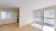 This thoughtfully designed single-floor residence offers a clean, modern aesthetic throughout. The open living area is anchored by light-toned hardwood flooring that extends seamlessly through the space, complemented by crisp white walls and recessed lighting. Large sliding glass doors flood the living room with natural light and provide direct access to a private balcony, offering views of the surrounding exterior corridor.

The galley-style kitchen is efficiently laid out with sage green cabinetry, white quartz countertops, a grid-pattern tile backsplash, and a speckled terrazzo floor. It is fully equipped with stainless steel appliances including a gas range, refrigerator, and microwave, alongside a deep undermount sink with a modern pull-down faucet.

The bedrooms are well-proportioned, each featuring the same warm wood flooring, horizontal windows that allow natural light, and roller shades for privacy. One bedroom includes a walk-in closet with built-in shelving and an accent green doorframe, while another is equipped with a wall-mounted mini-split air conditioning unit. The bathroom features full white square tile walls, a soaking tub with a glass panel, and brushed nickel fixtures. A shared outdoor corridor with open-air skylight access connects the unit to its surroundings.