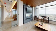 Located at 710 12th Street in Arlington, Virginia, this upscale amenity lounge occupies an elevated floor of a modern high-rise, offering an exceptional blend of contemporary design and comfort. The space is defined by expansive floor-to-ceiling glass curtain walls that flood the interior with natural light and frame sweeping panoramic views of the Arlington and greater Washington, D.C. metropolitan area.

Warm wood-slatted ceilings run throughout, complementing light hardwood flooring and creating a cohesive, refined aesthetic. Structural columns wrapped in matching wood veneer add architectural interest while maintaining an open, airy feel. The main lounge area features a sleek built-in fireplace beneath a wall-mounted television, set against a dramatic textured stone surround — a striking focal point for relaxed gathering.

Comfortable contemporary seating, including a gray sectional accented with vibrant orange and blue throw pillows, is arranged around low-profile wood coffee tables and artful décor. A counter-height bar table positioned directly against the glass offers an ideal perch to take in the sweeping outdoor views.

A dedicated dining area provides casual café-style seating with green chairs around white bistro tables, accompanied by pendant lighting and a textured accent wall with warm wood cabinetry. Glass doors open to an outdoor terrace, further connecting the interior to the impressive skyline surroundings. The overall ambiance is modern, polished, and welcoming.