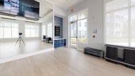 This single-floor fitness studio is thoughtfully designed to support a range of group exercise and barre-style classes in a clean, modern environment. The main exercise area is generously proportioned, featuring light wood-tone flooring that extends throughout, creating a warm yet polished aesthetic. Soft gray walls and crisp white trim contribute to the studio's airy, neutral palette.

A wall of floor-to-ceiling mirrors lines one side of the main studio, complete with a mounted wooden barre rail — a hallmark feature for barre and dance-fitness classes. A wall-mounted flat-screen display is positioned above the mirror wall, offering instructional or on-demand workout content. The studio benefits from an abundance of natural light, with large multi-pane windows on multiple walls offering views of lush palm trees and surrounding outdoor greenery.

Overhead, a coffered ceiling with suspended linear track lighting adds architectural interest while ensuring even illumination across the open floor. Step platforms and mat racks are stored neatly along the perimeter, keeping the main floor clear and functional.

The entryway area provides organized storage with a cubby unit holding folded towels and blue fabric bins, alongside wall-mounted foam roller racks. A glass-paned door connects the studio to an adjacent fitness area, enhancing the sense of openness and flow throughout the space.