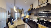 This well-designed single-floor residence offers a thoughtfully laid-out living space with a contemporary aesthetic throughout. The open-concept kitchen is a standout feature, showcasing rich dark cabinetry, light stone countertops, a mosaic tile backsplash, and a full suite of stainless steel appliances including a French door refrigerator, electric range, built-in microwave, and dishwasher. A peninsula counter with pendant lighting bridges the kitchen and living area, creating a seamless flow ideal for entertaining. The polished dark hardwood-style flooring extends throughout the main living spaces, adding a sleek, modern feel. The living area benefits from natural light provided by a glass door that opens to an exterior balcony, complemented by a ceiling fan overhead. The bedroom is carpeted for comfort and features a large window with decorative curtains, allowing ample natural light. Framed artwork above the bed and a cozy seating area contribute to an inviting atmosphere. The bathroom is equipped with a dark vanity topped with a light stone countertop, a wide mirror with overhead lighting, chevron-patterned tile flooring, and direct access to a spacious walk-in closet. Together, these spaces combine functional design with stylish finishes to create a cohesive and comfortable living environment.