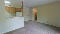 This single-floor residential space offers a practical and functional layout centered around an open-concept design. The kitchen is equipped with warm honey-toned wood cabinetry and a white refrigerator, and is defined by a peninsula counter with a decorative turned post detail, which separates it from the living area while maintaining an open, connected feel. The peninsula provides additional counter space and creates a natural transition between the two areas. The living area features wall-to-wall carpeting in a neutral tone, offering a soft and comfortable underfoot feel throughout. The walls are finished in a light beige palette, contributing to a bright and understated ambiance. A doorway leads from the main living space into an additional room, suggesting a functional corridor connecting the living areas to private spaces beyond. A closet door is also visible, providing built-in storage. Ventilation is handled via ceiling-mounted air vents, and the overall layout makes efficient use of the available space. The neutral color scheme and open flow between the kitchen and living room create a cohesive, easy-to-furnish environment well-suited for everyday living.