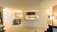 This single-floor residence offers an open-concept layout designed for comfortable everyday living. The kitchen is defined by a raised breakfast bar that serves as a natural divider between the cooking and living spaces, providing both functionality and visual separation without closing off the floor plan. Light-toned cabinetry, a white appliance suite, and an overhead microwave keep the kitchen feel clean and organized. The pass-through design allows for easy interaction between the kitchen and the adjoining areas. The dining space sits just beyond, illuminated by a stylish pendant light that draws the eye and anchors the area with a warm, inviting glow. A decorative sunburst mirror on the neutral-toned wall adds an artistic focal point to the dining nook. Soft wall-to-wall carpeting flows throughout the living and dining zones, contributing to the overall warmth and comfort of the interior. The neutral color palette — featuring soft beige and cream tones on both walls and flooring — creates a versatile backdrop that feels open and airy. The seamless flow between the kitchen, dining, and living areas makes the space well-suited for both relaxed daily living and casual entertaining.
