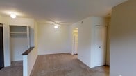 This single-floor space presents a clean, straightforward layout centered around a carpeted living area with neutral-toned walls that create a bright, understated ambiance. The room is equipped with a ceiling fan, providing both light and air circulation. The open sightline from the living area leads directly to an adjacent kitchen, visible through a doorway that reveals a full-sized refrigerator, suggesting a functional and accessible kitchen layout. A partial wall divides the living space from the kitchen area, adding a degree of visual separation while maintaining an open feel. To the right, a closet with a door offers convenient storage within the living area. A HVAC vent is positioned near the ceiling, indicating climate control throughout the unit. The flooring throughout the living area consists of beige carpet, contributing to a modest and comfortable atmosphere. Overall, the space is practical and straightforward, with a simple design that emphasizes functionality and ease of use. The light-colored walls and ceiling help to maximize the sense of brightness in the room.