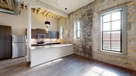 This single-floor loft masterfully blends historic industrial character with contemporary finishes. The open-concept living and kitchen area commands attention with soaring ceilings that expose original timber beam framing, creating a dramatic and airy atmosphere. Original exposed brick walls — worn to a rich, textured patina — run throughout the space, serving as a defining architectural feature that anchors the industrial aesthetic.

The gourmet kitchen is a standout, outfitted with dark wood cabinetry, stainless steel appliances including a built-in microwave, electric range, and full-size refrigerator, all complemented by striking marble-veined granite countertops. A peninsula with a built-in sink and elegant pendant lighting provides both functional workspace and a natural gathering point. The kitchen benefits from abundant natural light streaming through multiple black-framed windows.

The bedroom features soft carpeting underfoot, exposed brick walls, and an arched window that frames urban skyline views, lending the room both warmth and character. The bathroom is clean and functional, finished with wood-tone flooring and a glass-enclosed shower. Wide-plank hardwood-style flooring flows through the main living areas, tying together the modern and historic elements of this thoughtfully designed space.