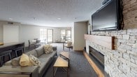 This single-level community room is thoughtfully designed to serve as a versatile gathering space for residents and guests. The main lounge area is anchored by a striking stacked-stone fireplace with a rich wood mantel and hearth, complemented by a wall-mounted flat-screen television — creating a warm and inviting focal point. Comfortable lounge seating, including a plush sofa and accent chairs, is arranged around a coffee table to encourage relaxed conversation and socializing.

The open layout seamlessly connects the lounge to multiple dining areas, each furnished with tables and chairs to accommodate various group sizes. A large kitchen bar with counter-height seating serves as a natural transition between the social and functional areas of the room. Natural light filters in through a set of glass-panel doors that open toward an outdoor area, brightening the space and enhancing its welcoming atmosphere.

The kitchen area is fully equipped with stainless steel appliances, including a full-size refrigerator and electric range, as well as a countertop workspace and a wall-mounted paper towel dispenser — making it well-suited for catering events or communal meals. Durable, wood-look flooring grounds the kitchen zone, while plush carpet throughout the main lounge and dining areas adds warmth and comfort. The textured ceiling and recessed lighting complete the polished yet approachable character of the space.