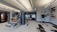 Located at 122 Parsons Ave in Columbus, Ohio, this well-appointed fitness center delivers a comprehensive workout environment designed for both casual exercisers and dedicated athletes. The single-floor facility is thoughtfully laid out to maximize usable space while maintaining an open, uncluttered feel throughout.

The cardio zone is positioned along a wall of large, floor-to-ceiling windows that flood the space with natural light and provide views of the surrounding area. Multiple treadmills and elliptical machines are arranged to take full advantage of this bright, energizing atmosphere. Elevated transom windows above the main panes further enhance the sense of openness and airiness.

The strength training area features a multi-station cable machine by TRUE Fitness, a variety of benches, and a well-stocked dumbbell rack spanning a broad weight range. Full-length mirrors line one wall, supporting proper form and creating a sense of added depth. Additional free-standing strength equipment, including a pull-up and dip station, rounds out the resistance training options.

Throughout the space, the design palette of cool gray walls, neutral carpet flooring, and exposed industrial ductwork strikes a balance between modern aesthetics and functional utility. Vintage-style fitness photography displayed along the walls adds a distinctive, motivating character. Restroom access is available on-site, and the facility is served by bright overhead linear lighting for consistent illumination at all hours.