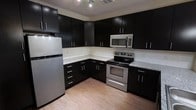 This single-floor kitchen showcases a contemporary design with bold, dark espresso-toned cabinetry that extends to the upper and lower sections, creating a sleek, cohesive aesthetic. The cabinetry is finished with brushed silver hardware, adding a subtle contrast against the deep tones. Light granite-style countertops run along the L-shaped layout, offering generous prep space while visually balancing the darker elements throughout the room. Stainless steel appliances, including a refrigerator, electric range, and over-the-range microwave, lend a polished, modern feel. The light wood-toned flooring provides a warm contrast to the darker upper elements, contributing to an overall balanced ambiance. Recessed track lighting is mounted on the ceiling, ensuring the workspace is well-illuminated. A clean white tile backsplash lines the walls between the upper and lower cabinets, enhancing the brightness of the space. The overall design strikes a balance between functionality and style, making it a practical yet visually appealing kitchen suited for everyday use.