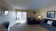 This single-floor space centers around a comfortable living room finished with wall-to-wall carpeting in a neutral tone, creating a soft, understated foundation throughout the area. A large sliding glass door dressed with vertical blinds serves as the room's primary source of natural light, allowing brightness to filter in while maintaining privacy. The door likely leads to an outdoor area, adding a sense of openness to the space. The walls are painted in a soft, warm beige, contributing to a calm and inviting atmosphere. A textured popcorn ceiling adds a classic character to the room. Climate control is provided by a wall-mounted air conditioning unit, positioned conveniently on the exterior wall. The living area offers ample room for seating arrangements, as evidenced by the space available for a sofa, loveseat, and accent chair. A small side table with a lamp provides warm ambient lighting in the corner, complementing the natural light from the sliding door. A decorative accent table near the entrance adds a personal touch. Overall, the space presents a functional and cozy layout well-suited for everyday living.