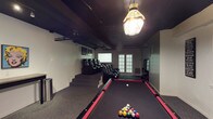 This single-floor entertainment space is thoughtfully designed to serve as a multipurpose recreational retreat. At the heart of the room sits a full-size pool table with a bold black felt surface and red trim, positioned beneath a classic stained-glass-style pendant light that casts a warm, inviting glow. The dark-painted ceiling enhances the intimate atmosphere, while exposed beams and overhead track lighting add a functional, industrial character to the space. Along one side of the room, a tiered platform elevates a row of plush black recliner chairs, creating a dedicated home theater-style viewing area ideal for movie nights or watching live sports. The projection screen mounted at the end of the room completes the cinematic setup. French doors along the rear wall introduce natural light and provide access to an adjoining area, balancing the room's otherwise moody ambiance. The dark carpeted flooring unifies the space and contributes to sound absorption, making it well-suited for both gaming and entertainment. A combination of recessed and overhead lighting ensures the room is well-lit throughout. Overall, this space offers a compelling blend of billiards, cinema, and lounge amenities within a single cohesive layout.