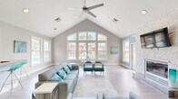 Located at 70 Greenway Square in Dover, Delaware, this single-floor community space is designed to impress with its dramatic vaulted ceilings and an oversized arched window wall that floods the interior with abundant natural light and offers views of the surrounding greenery and pool area. The open-plan layout comfortably accommodates multiple functional zones, all unified by light wood-look flooring and a crisp, neutral color palette accented with calming aqua tones. The main gathering area features a ceiling fan overhead and ample room for lounge seating, a glass-top dining table, and accent chairs, creating a welcoming social environment. A standout white stacked-stone fireplace anchors one wall, paired with a wall-mounted television, while a dedicated workspace with a built-in desk adds practical utility. Vibrant abstract artwork punctuates the walls throughout, contributing to the space's contemporary aesthetic. The fully equipped kitchen includes stainless steel appliances, white cabinetry, stone-look countertops, and a peninsula island, providing everything needed for community gatherings. A restroom is conveniently accessible via a connecting hallway. The overall ambiance is airy, modern, and inviting — ideal for community events and everyday use.
