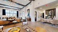 This expansive single-floor amenity facility offers a thoughtfully designed collection of spaces that blend contemporary style with practical functionality. The grand lobby lounge serves as the centerpiece, boasting soaring ceilings with bold dark beam accents, whitewashed brick columns, herringbone-patterned flooring, and a striking sputnik chandelier that anchors the open layout. Large windows flood the space with natural light, while curated furnishings in warm gold, leather, and wood tones create an inviting, upscale atmosphere.

Adjacent to the lounge is a dedicated leasing or reception area, featuring a comfortable seating arrangement, bold artwork, and a built-in workstation with marble-top cabinetry. A fully equipped café bar with warm wood cabinetry, white quartz countertops, pendant lighting, and a Starbucks coffee station adds a premium hospitality touch.

The game room provides a generous open area with wood-look flooring, a billiard table, a wall-mounted TV, and direct access to outdoor views through glass French doors. A private conference room with a large table, leather chairs, designer pendant lighting, and full-length mirrors offers a sophisticated meeting environment. A compact but well-appointed business center workstation with marble countertops and gold accent mirrors rounds out the work-focused amenities. The fitness center features a wide array of cardio and strength equipment alongside a mirrored wall and oversized windows with outdoor views.