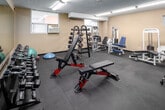This fitness facility at 100 Ferguson Ave S in Hamilton, Ontario, offers a well-organized, single-floor space designed to support a full range of workout activities. The interior features neutral beige walls, textured ceilings, and durable rubber flooring throughout, creating a functional and practical environment suited for regular use. Fluorescent lighting illuminates the space evenly, while small windows along the walls allow modest natural light to supplement the overhead fixtures.

The facility is divided into distinct exercise zones. The cardio area is well-stocked with multiple elliptical trainers, stationary bikes, treadmills, and a rowing machine, providing ample options for aerobic training. A wall-mounted television with a cable subscription is positioned for viewing during workouts. The strength training zone features a cable machine, weight rack with barbells and dumbbells, a squat rack, adjustable benches, and various resistance machines targeting major muscle groups. A large wall mirror in the strength area enhances spatial awareness and supports proper form during exercise.

Additional storage is provided via a wooden cabinet, and the space includes a dedicated area with a medicine ball and balance equipment for functional training. The overall layout is efficient and accessible, making this facility a comprehensive option for residents seeking convenient on-site fitness amenities.