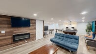 Located at 240 New State Rd in Manchester, CT, this thoughtfully designed recreational space offers a spacious, open-concept layout finished with light wood-look flooring, recessed lighting, and crisp white ceilings that keep the atmosphere bright and inviting. The space is anchored by a striking reclaimed wood accent wall, which hosts a wall-mounted flat-screen TV above a sleek electric fireplace — creating a warm and stylish entertainment focal point. Adjacent to this lounge area, comfortable futon-style seating is arranged around a wood coffee table, perfect for gathering and relaxing. A bold teal feature wall defines a secondary lounge zone, adding a vibrant pop of color and personality. The game area is well-equipped with a billiards table, a ping-pong table, and a full-length shuffleboard table, offering ample entertainment options. Pendant lighting illuminates a dual-workstation setup with monitors, making the space functional for productivity as well. Light blue-painted walls in the staircase area lend a calm, airy feel, while the open staircase with white railings provides clean, accessible flow. Throughout, the design balances modern aesthetics with recreational utility, making this a highly versatile and engaging space suited for leisure, socializing, or work.