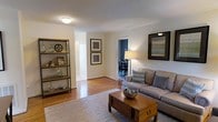 This single-floor residence offers a comfortable and cohesive living experience with a layout that flows naturally from room to room. The living area serves as the heart of the home, showcasing rich hardwood flooring that extends throughout the space and lends a warm, inviting ambiance. Neutral wall tones create a bright, airy atmosphere, complemented by a textured ceiling and flush lighting overhead.

The living room is generously proportioned, accommodating a plush sofa accented with decorative pillows, a rustic wooden coffee table, and an open metal shelving unit that adds an industrial touch while providing ample display and storage space. Framed artwork adorns the walls, contributing to the curated, homey feel of the space.

A well-lit dining area is visible through an open passageway, featuring a classic dining set bathed in warm lamplight against a deep accent wall that adds a striking contrast to the otherwise light palette. An additional hallway leads to further rooms, suggesting a practical and well-organized floor plan. The seamless connection between living, dining, and adjoining spaces makes this home ideal for both everyday living and entertaining.