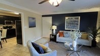 Located at The Flats At Hurstbourne, this single-story residence offers a thoughtfully designed living space that balances comfort with modern style. The living room serves as the focal point of the home, immediately drawing attention with a striking navy blue accent wall that creates a bold contrast against the warm, neutral-toned walls throughout. Crown molding adds an elegant architectural detail, lending a refined touch to the space.

The flooring transitions from light tile near the entryway to plush carpeting in the main seating area, providing both practicality and warmth underfoot. A ceiling fan with a decorative light fixture is centrally mounted, enhancing airflow and contributing to the room's overall comfort.

The layout accommodates a well-appointed seating arrangement, with ample room for lounge chairs and a glass-top coffee table. Thoughtful touches such as framed wall art, coordinated throw pillows in navy, orange, and geometric patterns, and layered area rugs contribute to a cohesive, inviting aesthetic. The white paneled entry door anchors the room's entryway, creating a clean and welcoming first impression throughout this well-maintained residence.