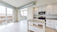 This two-bedroom, one-bathroom apartment at Hencen Apartments in Minneapolis, Minnesota offers a well-designed single-level layout with a clean, contemporary aesthetic throughout. The open-concept kitchen serves as a central highlight, featuring an abundance of white shaker-style cabinetry, gray countertops, and a full suite of stainless steel appliances, including a refrigerator, electric range, microwave, and dishwasher. A freestanding island with an open lower shelf provides additional workspace and storage. Light wood-tone flooring flows throughout the unit, lending warmth to the bright, neutral interior. Exposed silver ductwork runs along the ceiling, adding an industrial-modern touch to the living area, which connects directly to a private balcony through sliding glass doors. The bedrooms are generously sized, with large windows offering views of the surrounding Minneapolis streetscape, including the historic Aveda Institute building. A walk-in closet with wire shelving provides practical storage. The bathroom includes a full tub-shower combination, a white vanity, and matching light flooring. Recessed lighting and an abundance of natural light contribute to the apartment's fresh, airy ambiance.