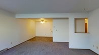 This single-story space presents an open layout divided into two distinct zones, differentiated by their flooring materials. The front area is finished with plush carpeting, creating a comfortable and casual atmosphere, while the adjoining room features dark tile flooring that adds a practical, easy-to-maintain surface. White walls throughout lend the space a clean, neutral backdrop, and the textured ceilings add a subtle layer of detail overhead. A ceiling fan with an integrated light fixture serves the tiled room, providing both ventilation and ambient lighting. A low partition wall with a pass-through opening connects the two areas, adding architectural interest while maintaining an open, flowing feel between spaces. The overall layout is straightforward and functional, with a simple, understated design that offers flexibility for various uses. The absence of clutter and the neutral color palette make the space feel airy and ready for personalization.