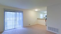This single-floor residence offers a clean, functional layout designed to maximize comfort and usability. The living area is bright and airy, featuring soft wall-to-wall carpeting and a large sliding glass door dressed with vertical blinds that floods the space with natural light while providing access to the outdoors. The neutral, light-toned walls lend a calm and welcoming ambiance throughout.

An open pass-through counter connects the living area to the kitchen, creating a sense of openness and ease of interaction between the two spaces. The kitchen is appointed with white cabinetry, a stainless steel microwave, and additional appliances, all illuminated by overhead lighting. A window in the kitchen brings in additional natural light, enhancing the bright and practical feel of the cooking area.

Ceiling-mounted flush lighting provides consistent illumination throughout the living space, complementing the natural light. A floor vent indicates a central HVAC system for year-round climate control. The overall design is straightforward and well-maintained, making it a versatile and move-in ready space suited for everyday living.