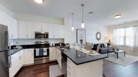 This single-floor space showcases a thoughtfully designed open-concept layout that connects the kitchen and living area in a cohesive, functional arrangement. The kitchen is outfitted with crisp white shaker-style cabinetry, dark granite countertops, and a full suite of stainless steel appliances, including a range, built-in microwave, and dishwasher. A central island extends the counter space and doubles as a dining area, illuminated overhead by a pair of modern pendant lights that add warmth and visual interest. Rich dark hardwood flooring runs throughout, providing a striking contrast against the white cabinetry and light-colored walls. The open flow into the living area allows for a generous sense of space, where a comfortable seating arrangement is visible, accented by decorative elements including a sunburst mirror and framed artwork. The overall aesthetic balances contemporary design with a welcoming ambiance, making the space ideal for both daily living and entertaining. Clean lines, neutral tones, and well-chosen fixtures contribute to a polished, modern feel throughout.