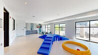 This well-appointed pet amenity facility spans a single floor and is thoughtfully divided into two distinct areas tailored for pets and their owners. The main play area features a durable speckled terrazzo-style floor, bright white subway tile walls, and large black-framed windows that flood the room with natural light and offer views of the surrounding neighborhood. Vibrant blue agility equipment, including ramps and tunnel structures, along with a yellow circular play ring, provide ample enrichment for dogs of all sizes. Colorful framed dog portraits add a playful, welcoming character to the walls. A cozy lounge nook with modern sculptural seating, a round table, and a wall-mounted TV creates a comfortable spot for pet owners to relax. The secondary room serves as a dedicated pet grooming station, featuring a built-in wash tub with step access, a grooming table with a mounted dryer, and striking teal accent tile that contrasts beautifully with the white subway tile walls. Recessed lighting throughout both areas ensures a clean, modern aesthetic, while a glass-paneled entry door connects the two spaces seamlessly. This facility offers a comprehensive, design-forward environment for pet care and recreation.