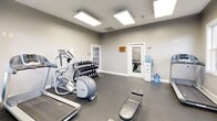 The fitness center at Campus Pointe in Charleston, IL, is a compact, well-equipped exercise space designed to support a range of workout routines. The room features neutral gray walls accented by a bold teal blue feature wall, creating an energetic yet focused atmosphere. Rubber flooring throughout provides durability and traction suitable for active use.

Natural light enters through multiple white-framed windows offering views of the surrounding greenery, complemented by recessed panel lighting in the ceiling that ensures consistent, bright illumination throughout the space. A large wall-mounted mirror enhances the sense of openness and allows users to monitor their form during exercise.

The fitness center is outfitted with a variety of cardio equipment, including treadmills, a Precor elliptical, and a stationary bike. A multi-functional cable machine serves as the centerpiece for strength training, accompanied by a full dumbbell rack and an adjustable weight bench. A rack of exercise and medicine balls in assorted sizes adds versatility for functional fitness routines. A water cooler is conveniently positioned near the entrance. The space is accessed through a classic paneled door and connects to an adjacent room, making it a practical and functional amenity.