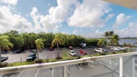 This single-floor space offers a notable outdoor balcony area that serves as one of its most distinctive features. The balcony is outfitted with a clean white railing and provides an elevated vantage point overlooking a well-maintained parking area framed by an abundance of tropical landscaping. Tall palm trees and dense green foliage create a lush, verdant backdrop, while a glimpse of open water is visible in the distance, lending the space a distinctly coastal Florida ambiance. The wide-open sky above, dotted with rolling clouds, enhances the sense of openness and connection to the outdoors. The bright, natural light characteristic of a sunny subtropical setting permeates the balcony, making it an inviting spot to take in the surrounding scenery. The interior portions of the space were not sufficiently illuminated in the available imagery to provide detailed observations; however, the overall setting and exterior presentation suggest a space situated within a multi-unit building that benefits greatly from its tropical surroundings and outdoor orientation. The combination of natural greenery, waterway views, and abundant sunlight gives this space a relaxed, resort-like character well-suited to the South Florida lifestyle.