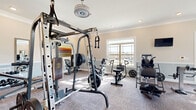 This fitness center offers a thoughtfully designed workout space that accommodates a wide range of exercise needs. The room features elegant crown molding and a two-tone wall treatment with soft neutral tones above and a muted blue-gray wainscoting below, lending the space a polished, welcoming ambiance. Durable patterned carpet covers the floor throughout, providing both comfort and stability underfoot. Large windows along multiple walls flood the interior with natural light and offer views of the surrounding neighborhood, creating an open and energizing atmosphere. The space is comprehensively outfitted with strength training equipment, including a multi-function cable and power rack system, a weight bench, and a dumbbell rack. Cardio options include treadmills, a stationary bike, an elliptical, a rowing machine, and an air bike. Full-length mirrors are positioned along certain walls to assist with form and create a sense of added depth. Mounted flat-screen televisions provide entertainment during workouts. Additional amenities include built-in cabinetry for storage, a wall-mounted drinking fountain, and a dedicated restroom. Recessed lighting and a ceiling fan round out the space, ensuring comfortable conditions during any workout session.