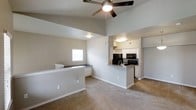 This single-story space showcases an open-concept design that seamlessly connects the living area to the kitchen. The living space is finished with wall-to-wall carpeting and features a ceiling fan mounted overhead, contributing to both comfort and airflow. The elevated ceiling height in the main living area adds a sense of openness and volume to the space.

The kitchen is accessible via a peninsula-style counter with a sink, providing a natural transition between the two areas. It is equipped with dark appliances, including a refrigerator, microwave, and range, which contrast with the white cabinetry above and below. The kitchen benefits from its own overhead lighting, keeping the workspace well-illuminated. A wine glass rack is mounted beneath the upper cabinets, adding a functional detail to the space.

Adjacent to the living area, a built-in counter or desk with white cabinetry and dark countertop is positioned near a window, offering a dedicated workspace or utility area. The neutral gray-toned walls throughout maintain a clean, cohesive aesthetic. Overall, the layout is practical and efficient, designed to maximize usability across the open floor plan.
