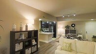 This single-floor residence offers a thoughtfully designed open-concept layout that seamlessly connects the living, dining, and kitchen areas into one cohesive and inviting space. The living area is anchored by warm, ambient lighting and features rich hardwood-style flooring that flows throughout, creating a sense of continuity and warmth. A stylish open shelving unit provides both storage and a decorative focal point, complementing the neutral, earthy tones of the walls.

The kitchen is a standout feature, boasting dark espresso cabinetry, stainless steel appliances — including a full-size refrigerator and microwave — and a peninsula with a light-colored countertop that doubles as a casual dining or prep space. Under-cabinet lighting adds a refined touch and enhances visibility in the cooking area.

Track lighting runs along the ceiling in the main living space, offering adjustable illumination that highlights the room's clean, modern aesthetic. The dining area is defined by a sleek glass-top table, adding an element of sophistication. Throughout the space, the design strikes a balance between contemporary style and comfortable livability, making it a functional and visually appealing environment.